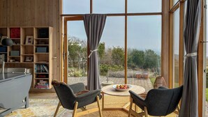 Una habitación con un ventanal grande, una estantería de madera, dos sillas, una mesa redonda con fruta y una alfombra.