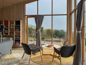 Ein Zimmer mit einem großen Fenster, einem hölzernen Bücherregal, zwei Stühlen, einem runden Tisch mit Obst und einem Teppich.