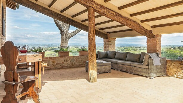 Eine Terrasse im Freien mit einem Holztisch, einem Korbsesselsofa und Blick auf die Landschaft.