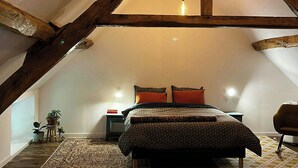 Quarto com teto inclinado, vigas de madeira, cama com roupa de cama estampada, mesa de cabeceira, uma cadeira e um lustre.