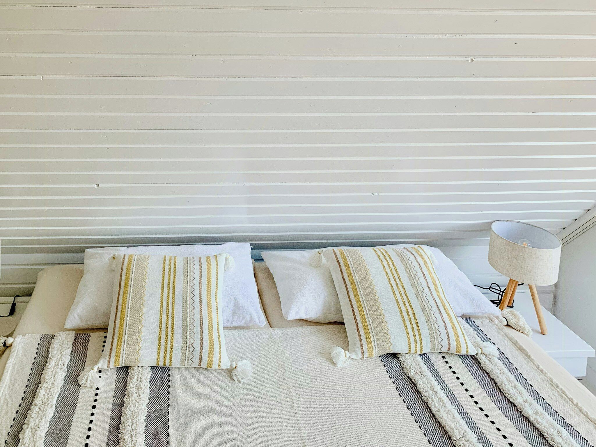 Una cama con almohadas a rayas, cabecera blanca, una lámpara de noche y una manta a rayas.
