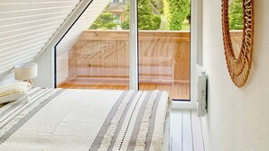 A bedroom with a sloped ceiling, a bed with a striped blanket, a bedside lamp, and a view of a wooden deck and garden through a sliding glass door. - Thatched holiday home by the Baltic Sea (Großenbrode)