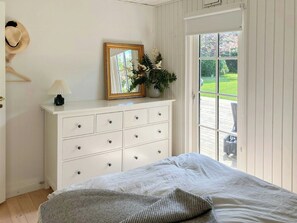Ein Schlafzimmer mit Bett, Kommode mit Schubladen, Spiegel, Lampe, Hut an der Wand und Blick ins Freie.