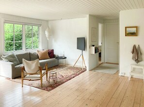 Ein Zimmer mit einer Couch, einem kleinen Tisch, einem Sessel, einem Fernseher auf einem Stativ, einem Teppich und einer Tür zu einem anderen Raum.
