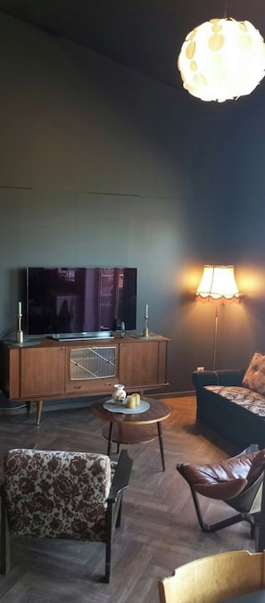 A living room with a flat-screen TV, a wooden cabinet, a round table with a lamp, and patterned chairs. - Renovated chapel with fjord views in Vågstranda (Vågstranda)