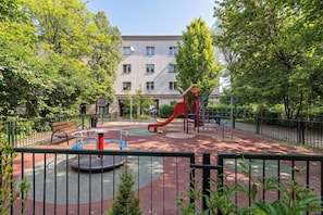A playground with a red slide and a merry-go-round, surrounded by trees and a building in the background. - Cozy Apartment | Near the Banksy Museum (Kraków)