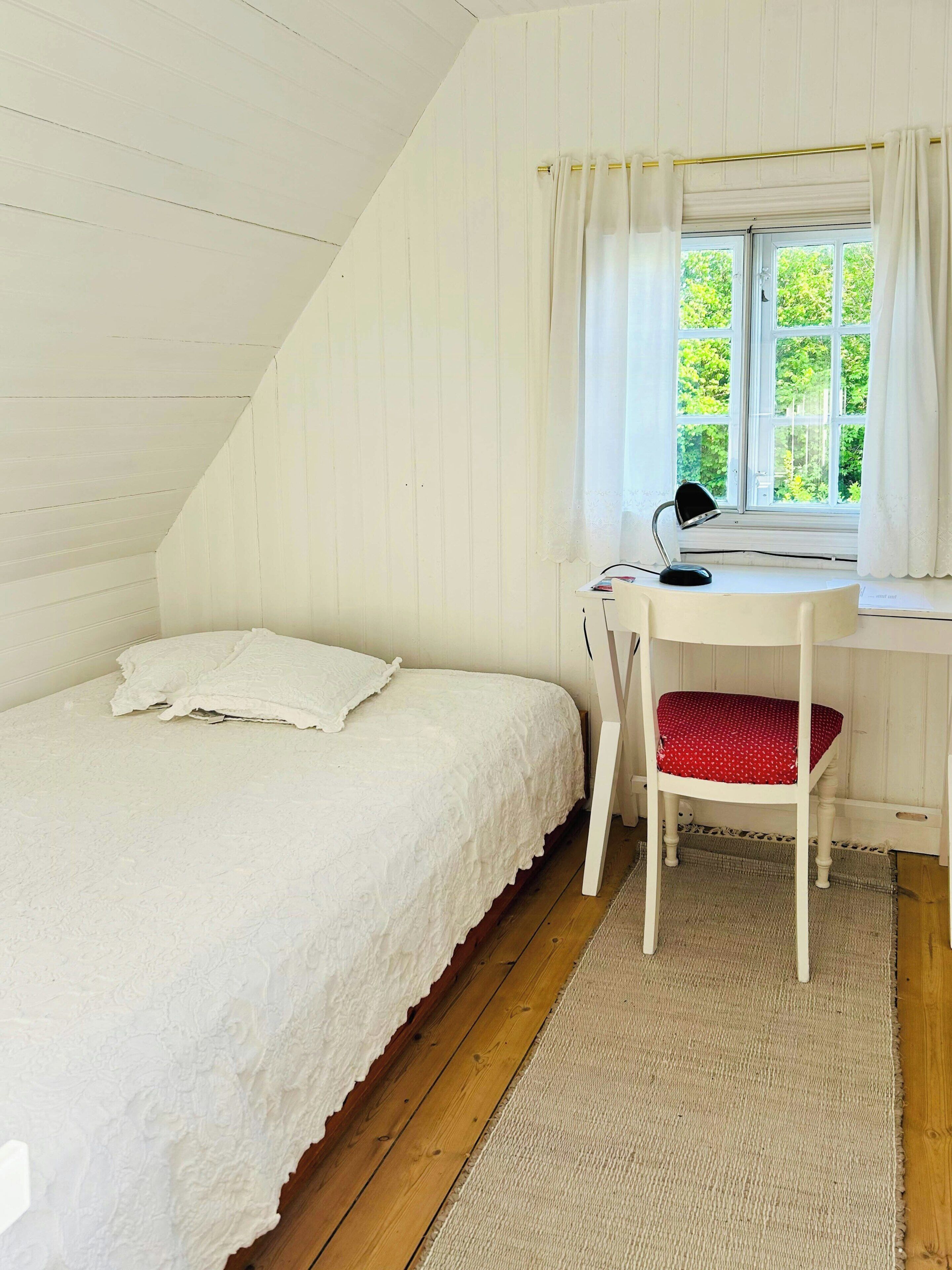 A bedroom with a bed, a desk with a chair, a window with curtains, and a view of greenery outside.