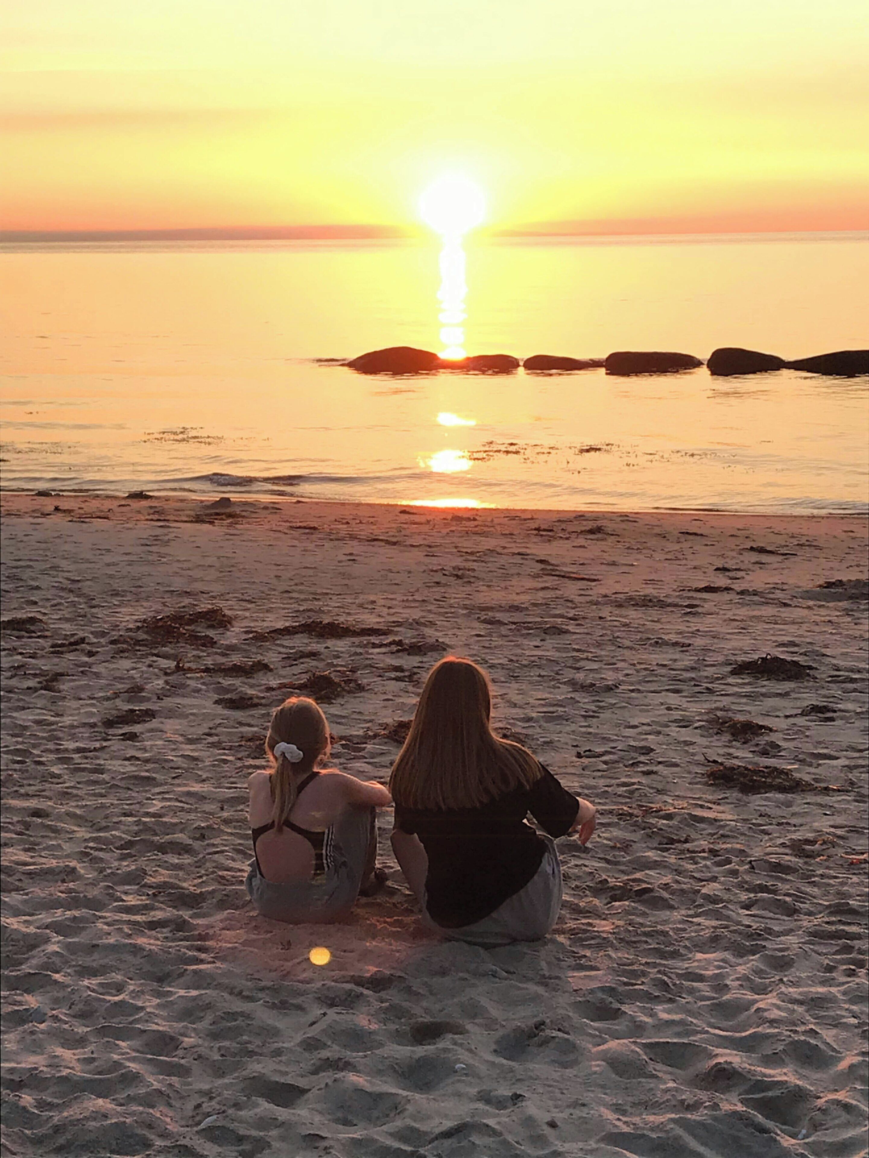 Two people sitting on a sandy beach facing the ocean during sunset.