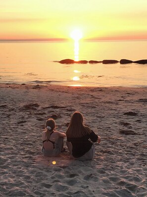 Zwei Personen sitzen am sandigen Strand und blicken auf den Ozean bei Sonnenuntergang.