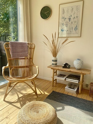 Ein Zimmer mit einem Korbstuhl, einer hölzernen Bank mit Büchern, einer Vase mit Trockenpflanzen und einem gerahmten Pflanzenblatt-Aufsatz an der Wand.
