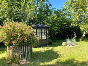 Ein Garten mit einem Holzschuppen, einem Strauch mit rosafarbenen Blüten und einer Rasenfläche.