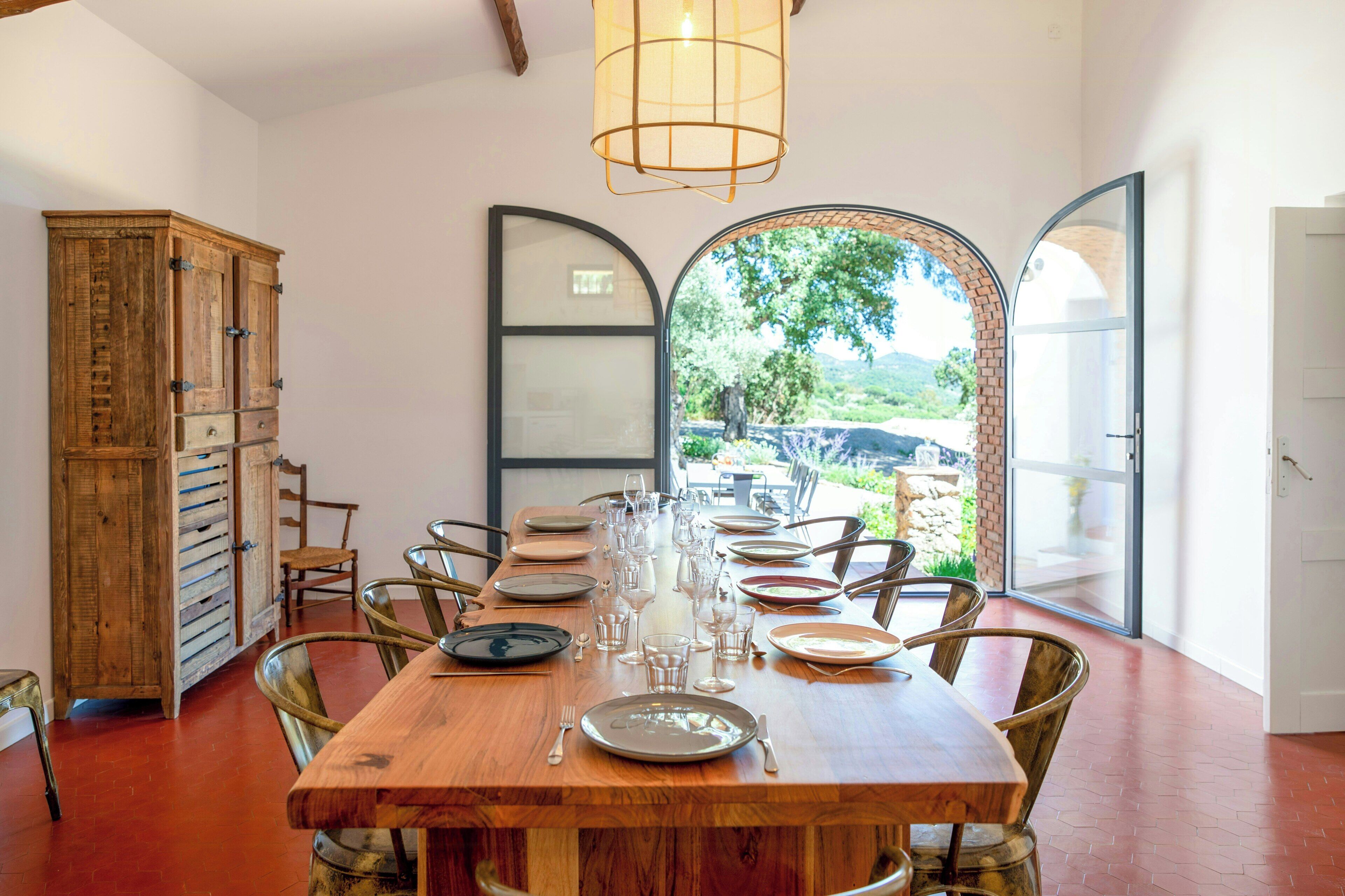 A dining area with a wooden table set for six, surrounded by chairs, a cabinet, and a large arched window offering a view of greenery.