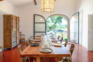 A dining area with a wooden table set for six, surrounded by chairs, a cabinet, and a large arched window offering a view of greenery. - 17th century Provençal farmhouse with pool (Le Plan de la Tour)