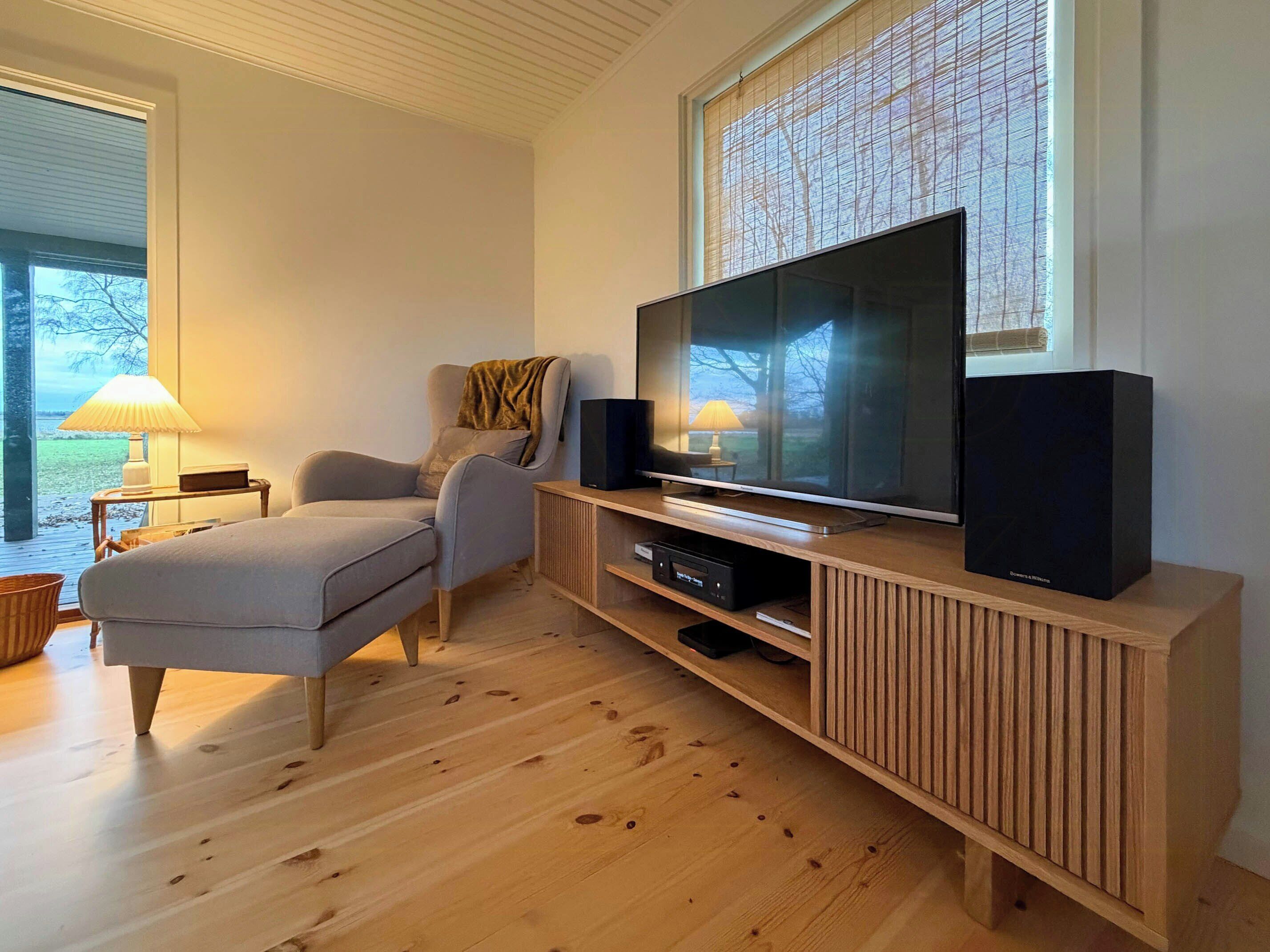 Een moderne woonkamer met een flatscreen televisie, een houten tv-meubel, een grijze bank en een raam met jaloezieën.