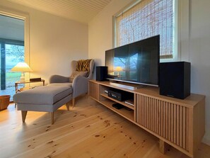 A modern living room with a flat-screen TV, wooden entertainment unit, grey sofa, and a window with blinds. - Stay very close to nature and the sea (Jægerspris)
