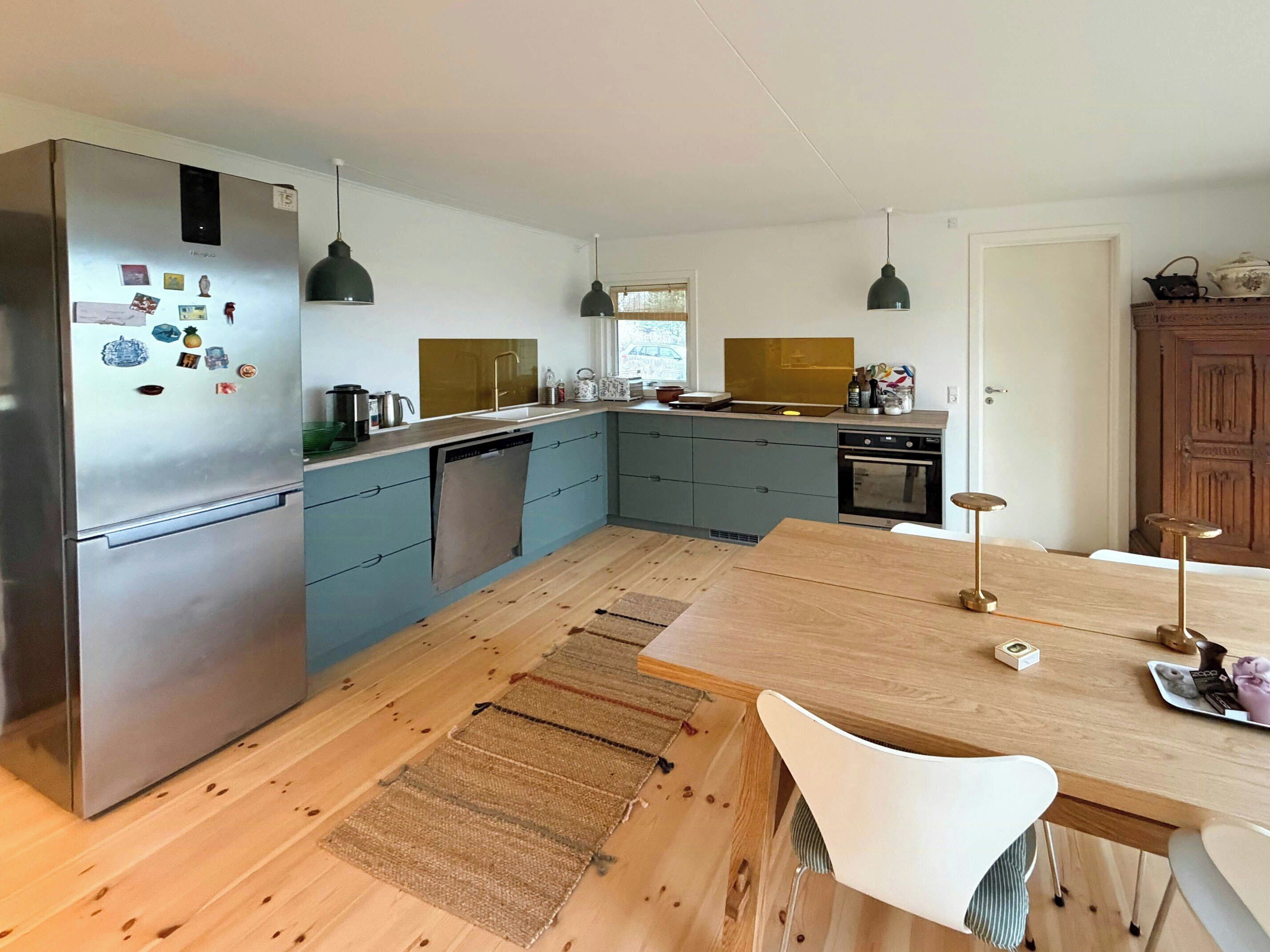 A modern kitchen with a stainless steel refrigerator, a dishwasher, a sink, and a wooden dining table with chairs.