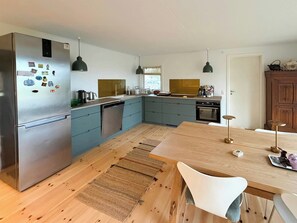 A modern kitchen with a stainless steel refrigerator, a dishwasher, a sink, and a wooden dining table with chairs. - Stay very close to nature and the sea (Jægerspris)
