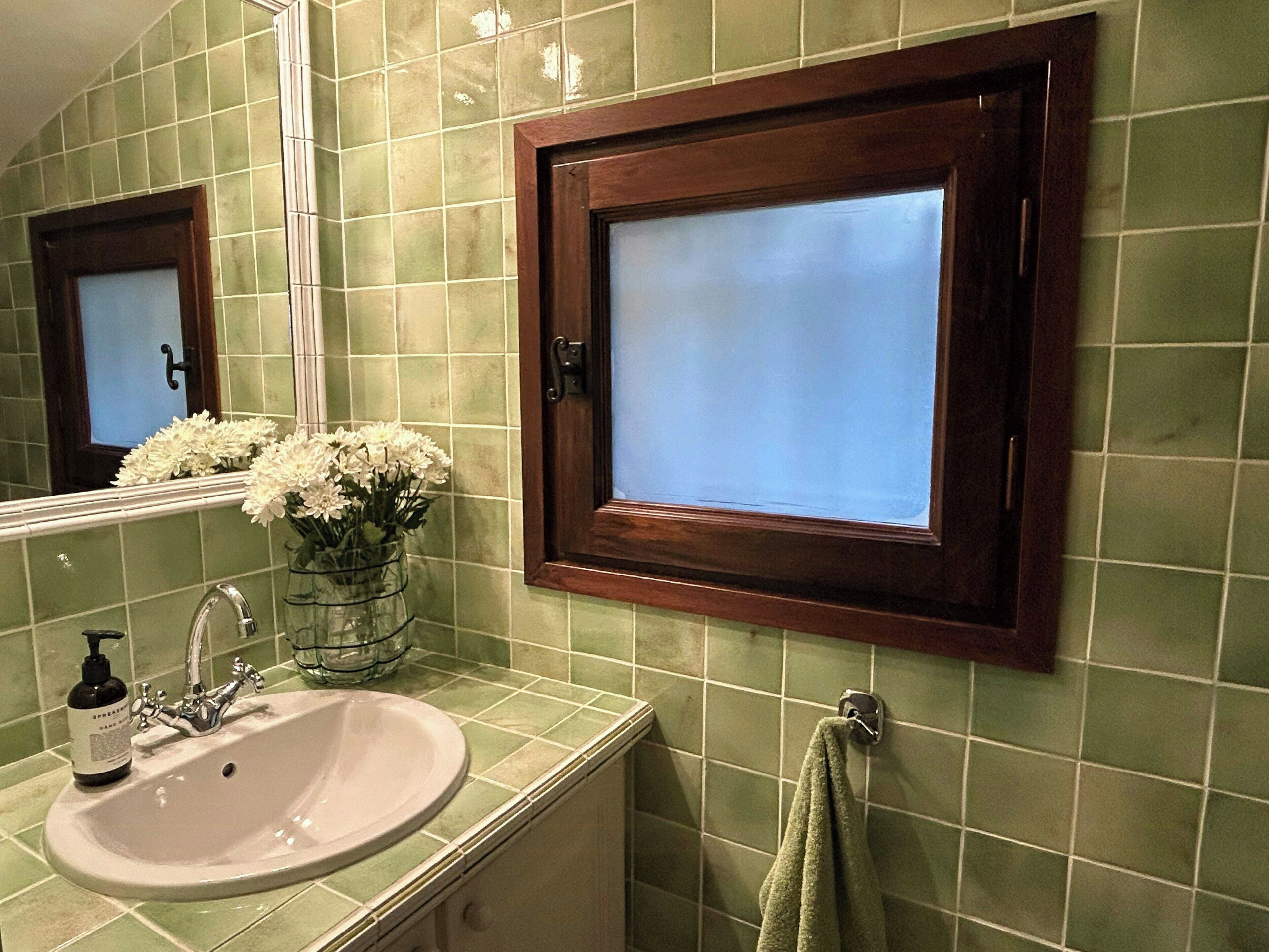A bathroom with a green tiled wall, a wooden-framed mirror, a sink with a faucet, a vase with flowers, and a towel hanging on a rack.