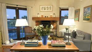Un salon avec une cheminée, un canapé, une table à manger avec des livres, et une vue dégagée grâce à de grandes fenêtres.