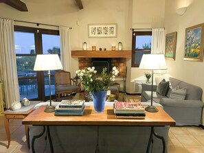 Ein Wohnzimmer mit Kamin, einer Couch, einem Esstisch mit Büchern und Blick durch große Fenster.