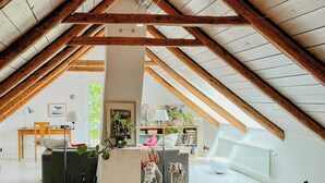 Une chambre mansardée avec un plafond haut, des poutres en bois, un bureau, une chaise, un canapé, un tapis et un chien.