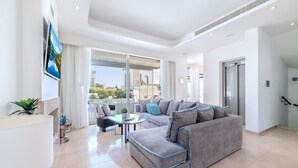 A modern living room with a grey sofa, a coffee table, and a large window with a view of greenery. - Lombardi Villa Protaras (Protaras)