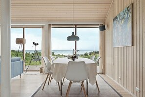 A dining area with a round table, white chairs, a hanging light fixture, and a view of the sea. - Seaside cottage at Bønnerup Strand (Glesborg)