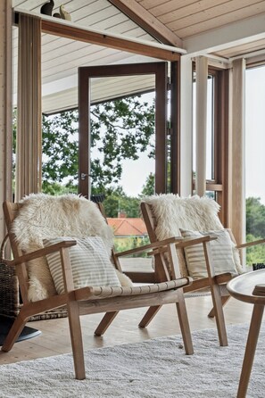A wooden chair with a fur throw and a cushion, a round table, and a view of trees through large windows. - Seaside cottage at Bønnerup Strand (Glesborg)