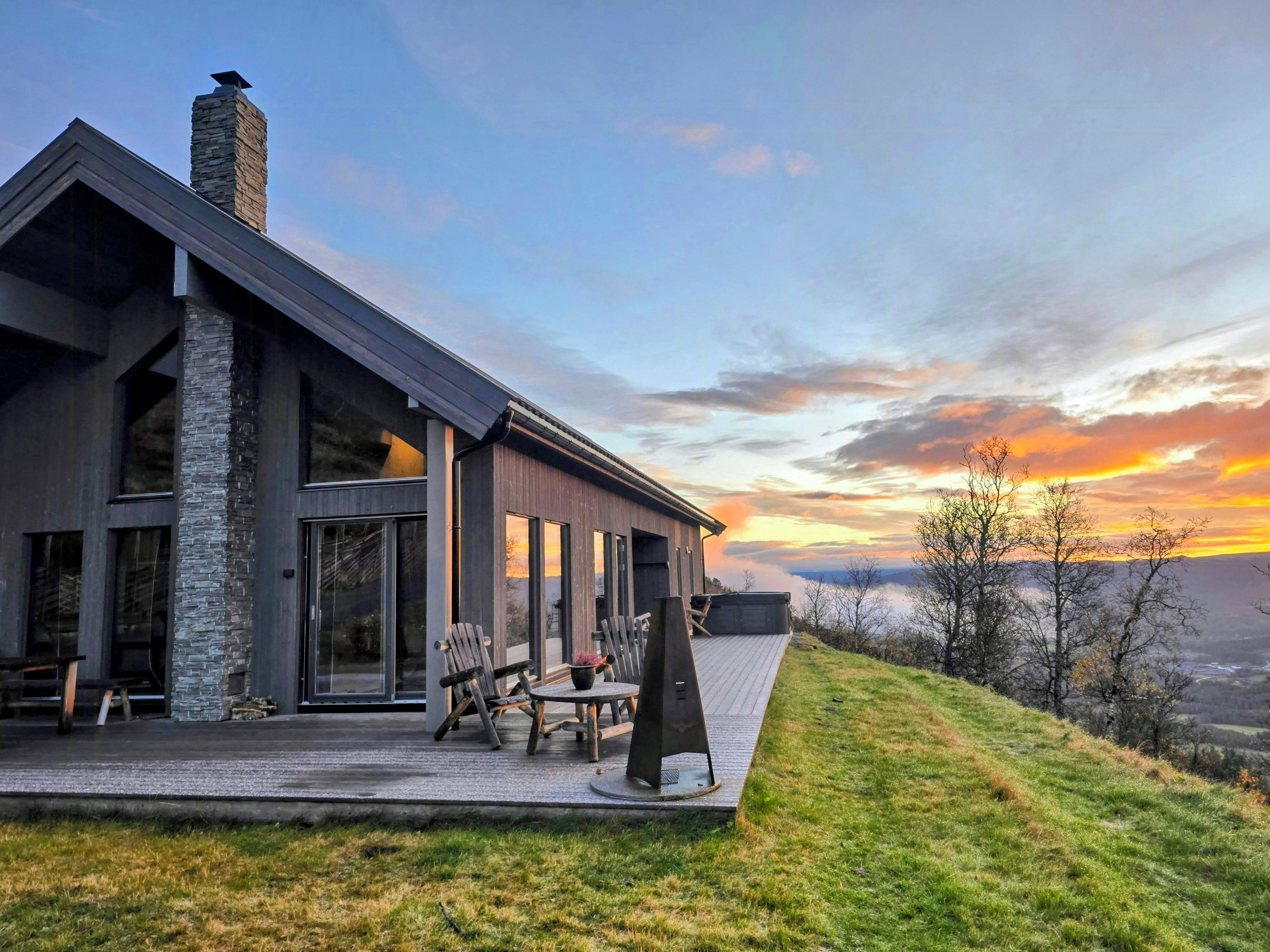 Une maison moderne avec une cheminée en pierre, de grandes fenêtres et une terrasse aménagée avec du mobilier d’extérieur.