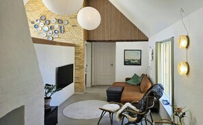 Ett vardagsrum med en tegelvägg, en platt-TV, en brun soffa, flätade stolar, en rund matta och en dörr som leder till ett annat rum.