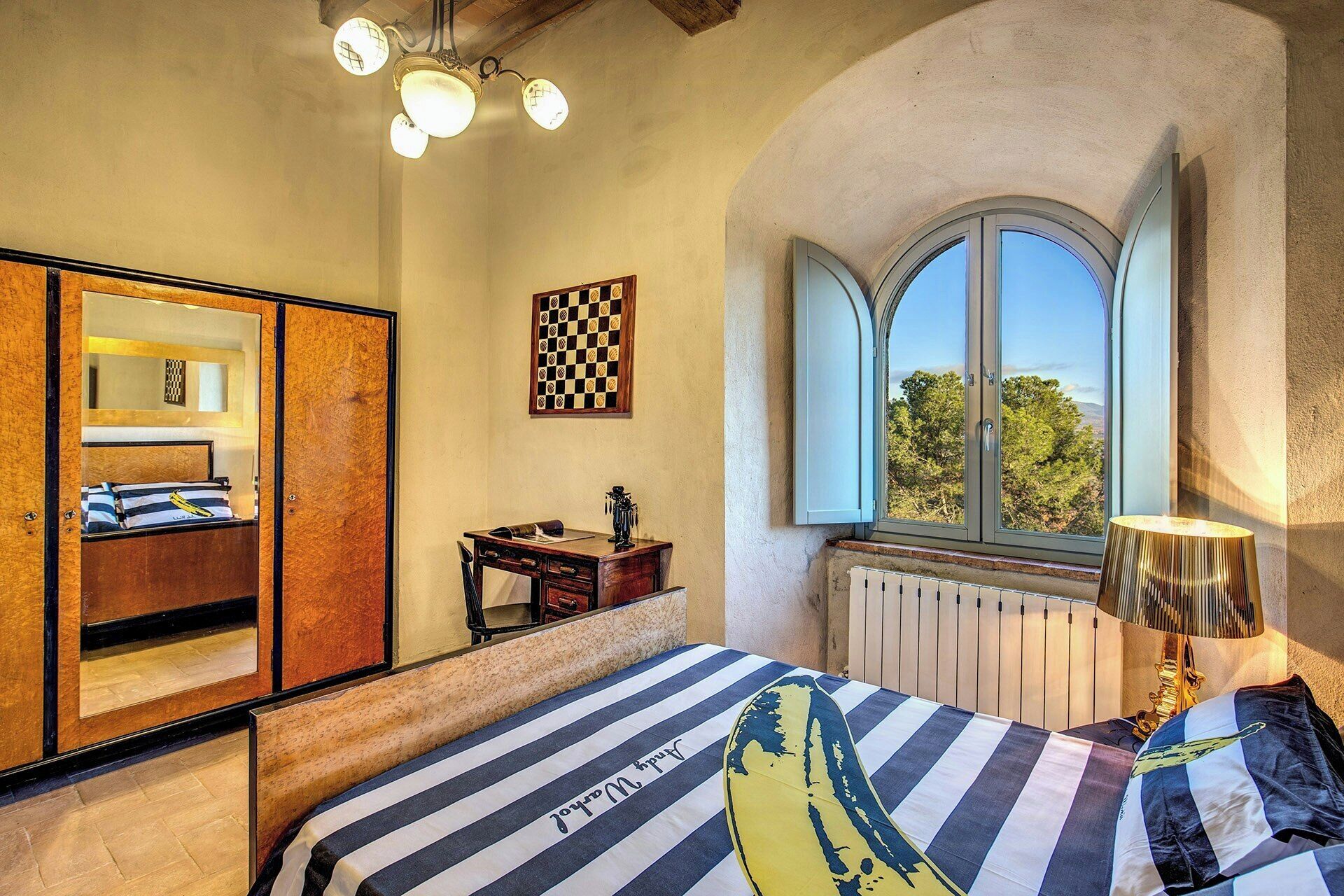 A bedroom with a bed, a mirror, a chessboard, a window, and a lamp.