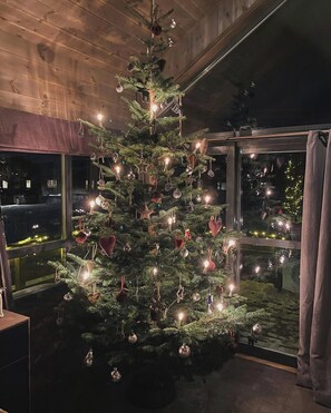 A Christmas tree with lights and ornaments, positioned in a room with wooden walls and large windows. - Family cabin with views of Store and Lille Hødn (Hemsedal)