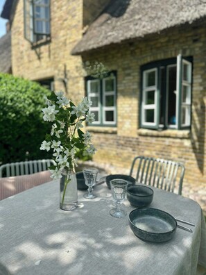Ein Tisch draußen gedeckt, mit einem Blumenstrauß, einer Schüssel und einem Teller, vor einem Gebäude aus Stein mit Fenstern.