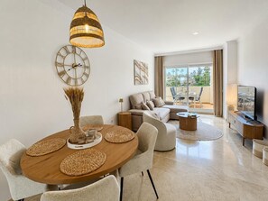 Ein modernes Wohnzimmer mit einem runden Holzesstisch, beigen Sofas, einem Fernseher und einem großen Fenster mit Blick auf Palmen.