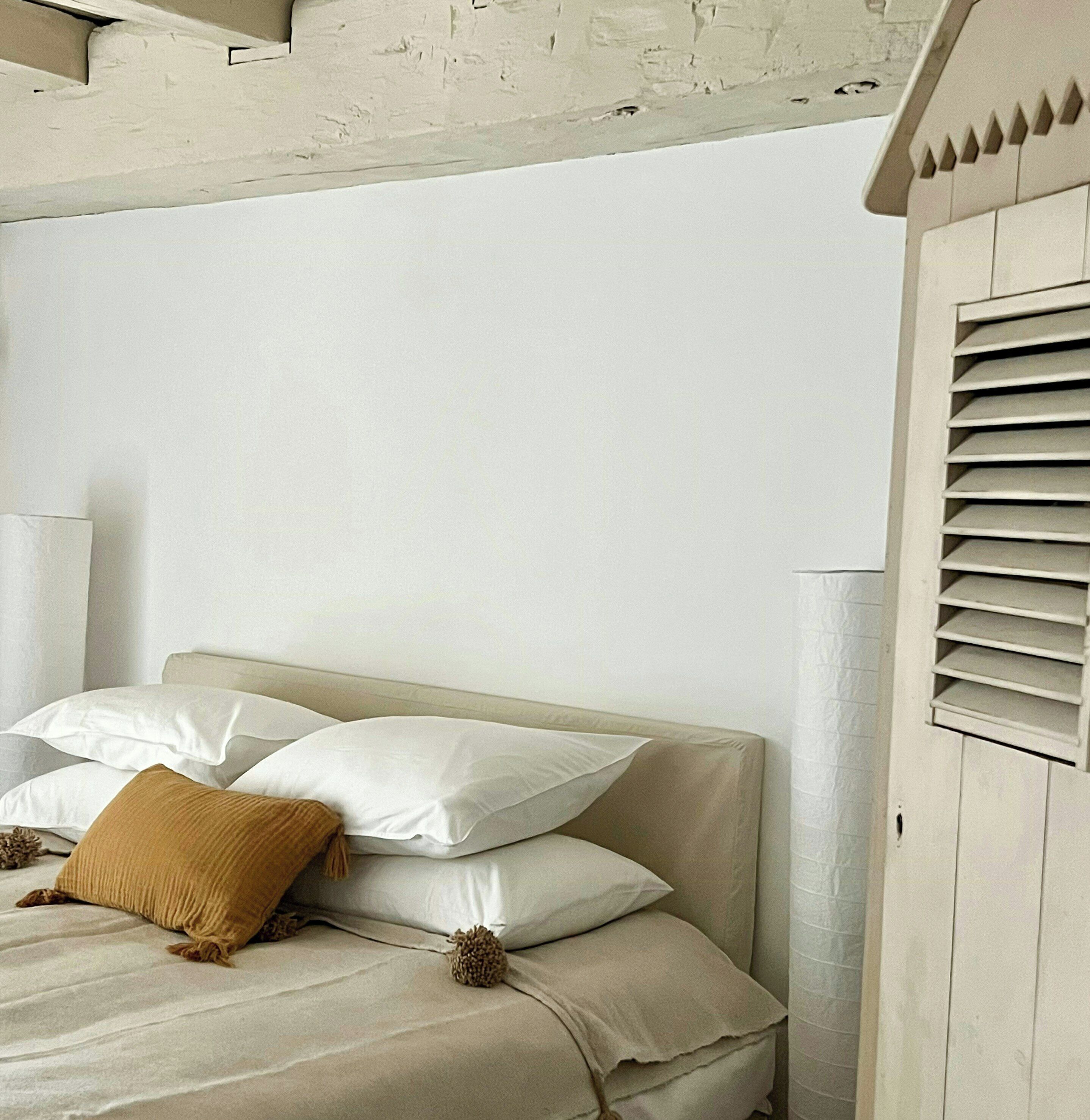 Ein Bett mit weißen Leinentüchern und einem braunen Kopfkissen, eine weiße Wand und ein Holz-Kopfteil.