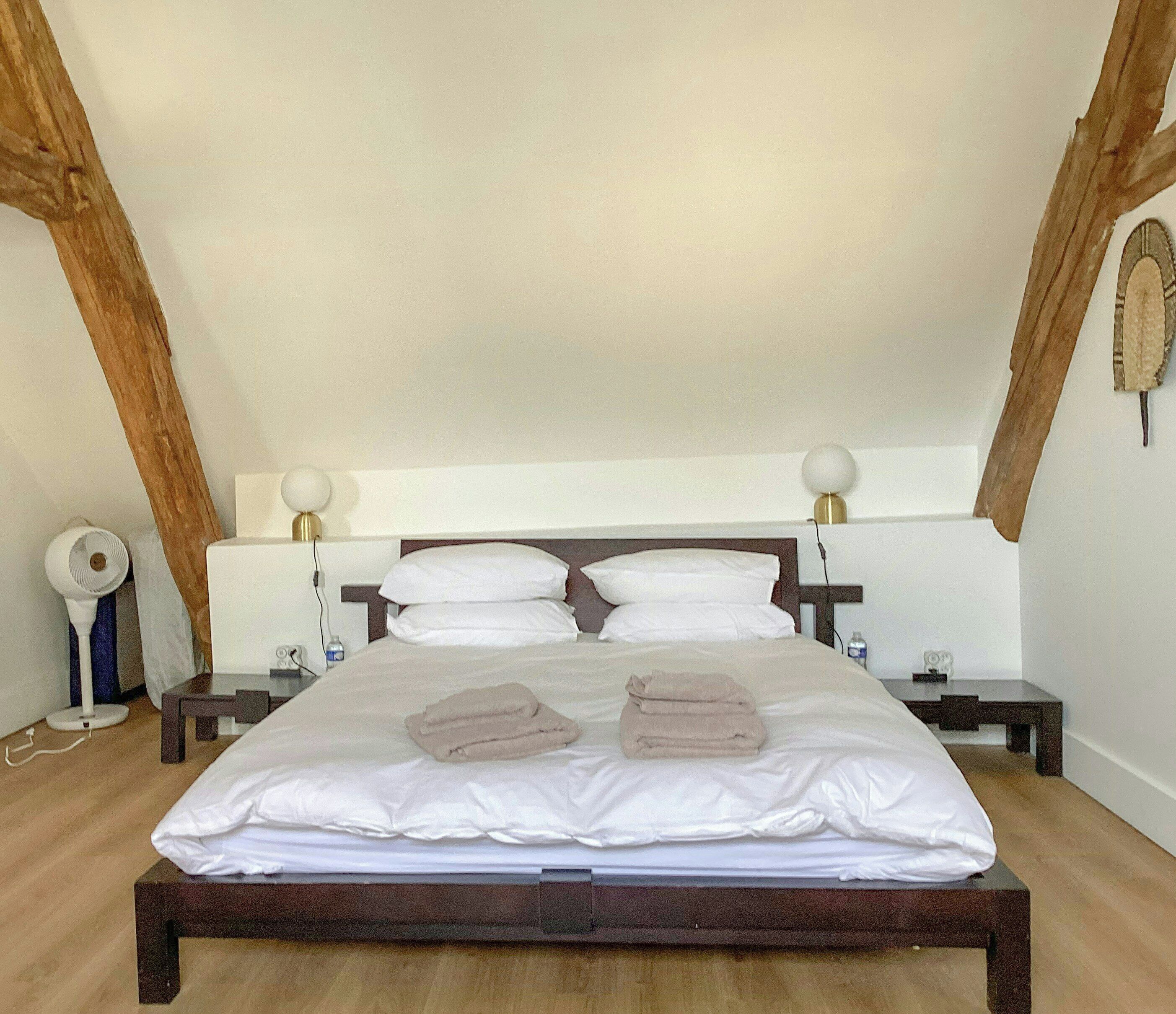 A neatly made bed with white linens and two folded towels, a bedside table with a lamp, and a wall-mounted fan.