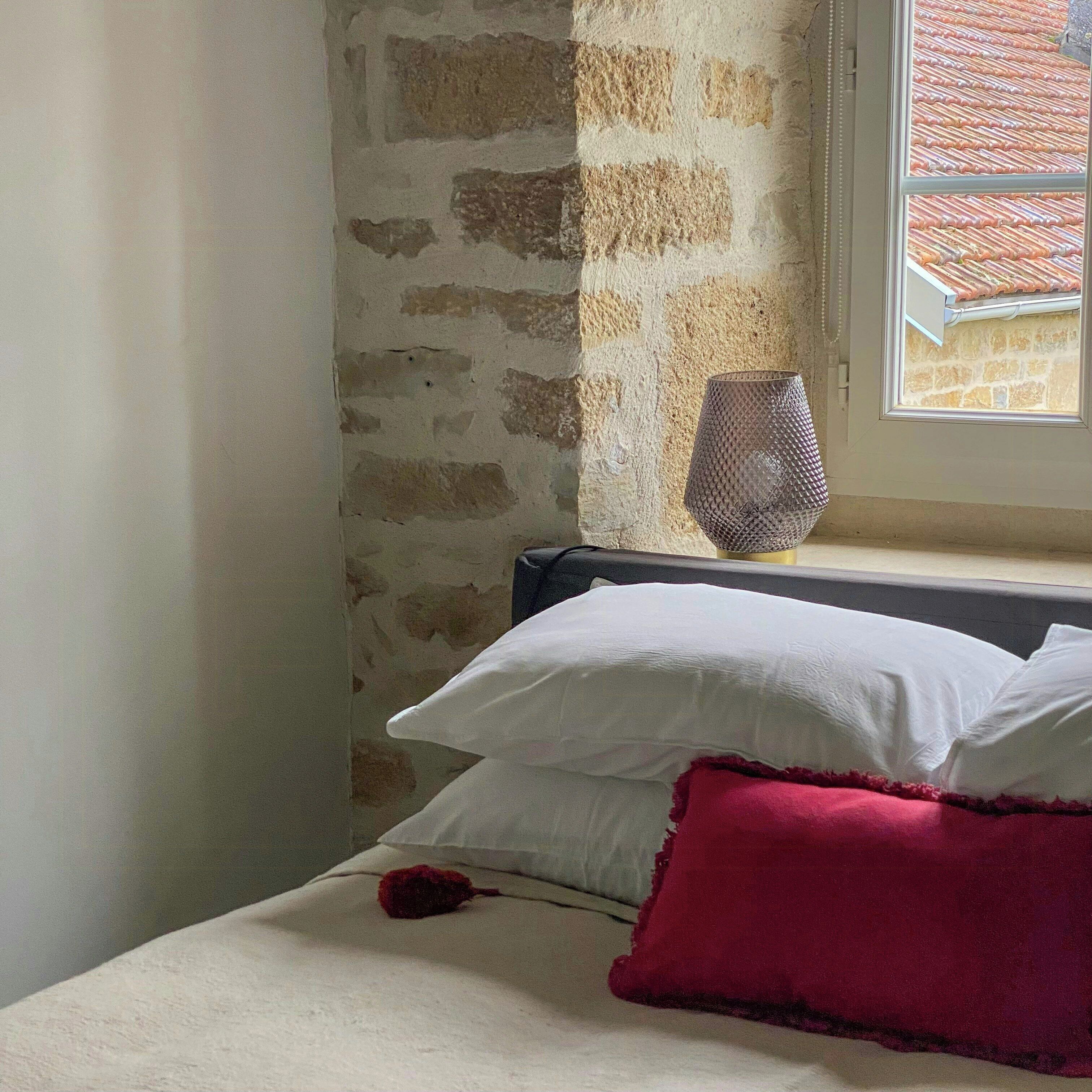 Et soveværelse med en stenvæg, et vindue med udsigt til et tegltag, en senget lampe og en seng med puder.