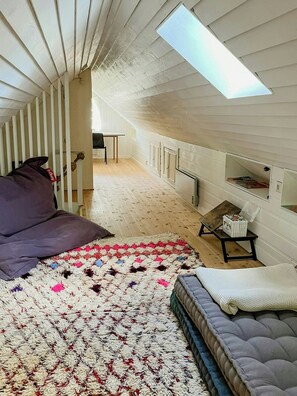 Ein gemütliches Schlafzimmer im Loft-Stil mit hohen Decken, einem Oberlicht, einem Bett mit farbenfroher Decke, einem kleinen Tisch und einem Sessel.