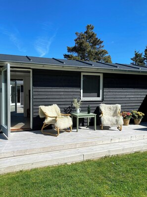 Ein Haus mit überdachter Terrasse, einem weißen, flauschigen Sessel, einem kleinen Tisch und Topfpflanzen.