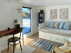 Ein gemütliches Zimmer mit einem Holztisch, einem weißen Sofa mit gemusterten Kissen, einem Bücherregal und gerahmten Pflanzenbildern an der Wand.