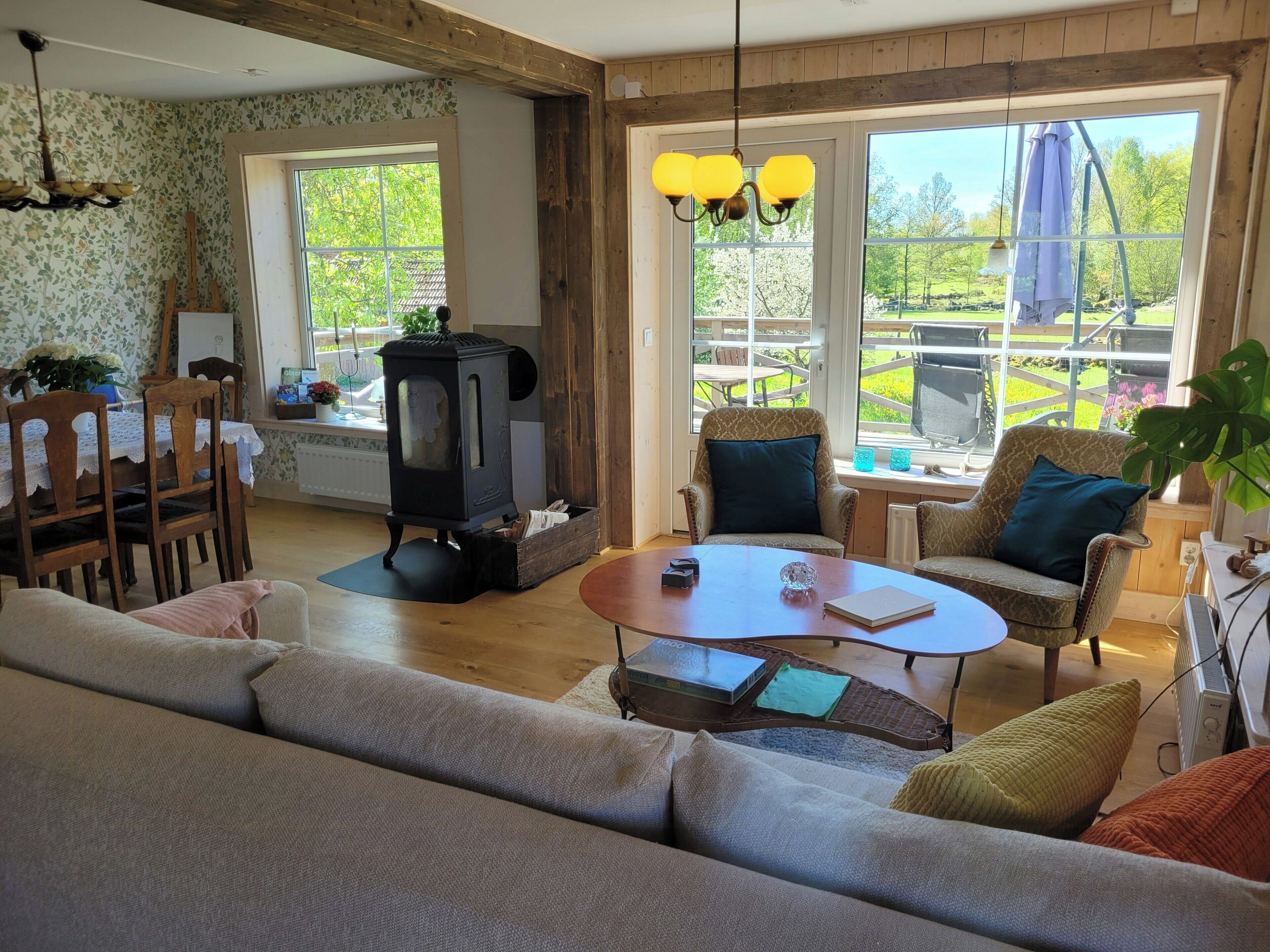 A cozy living room with a sofa, chairs, a table, a fireplace, and large windows with a view of greenery.