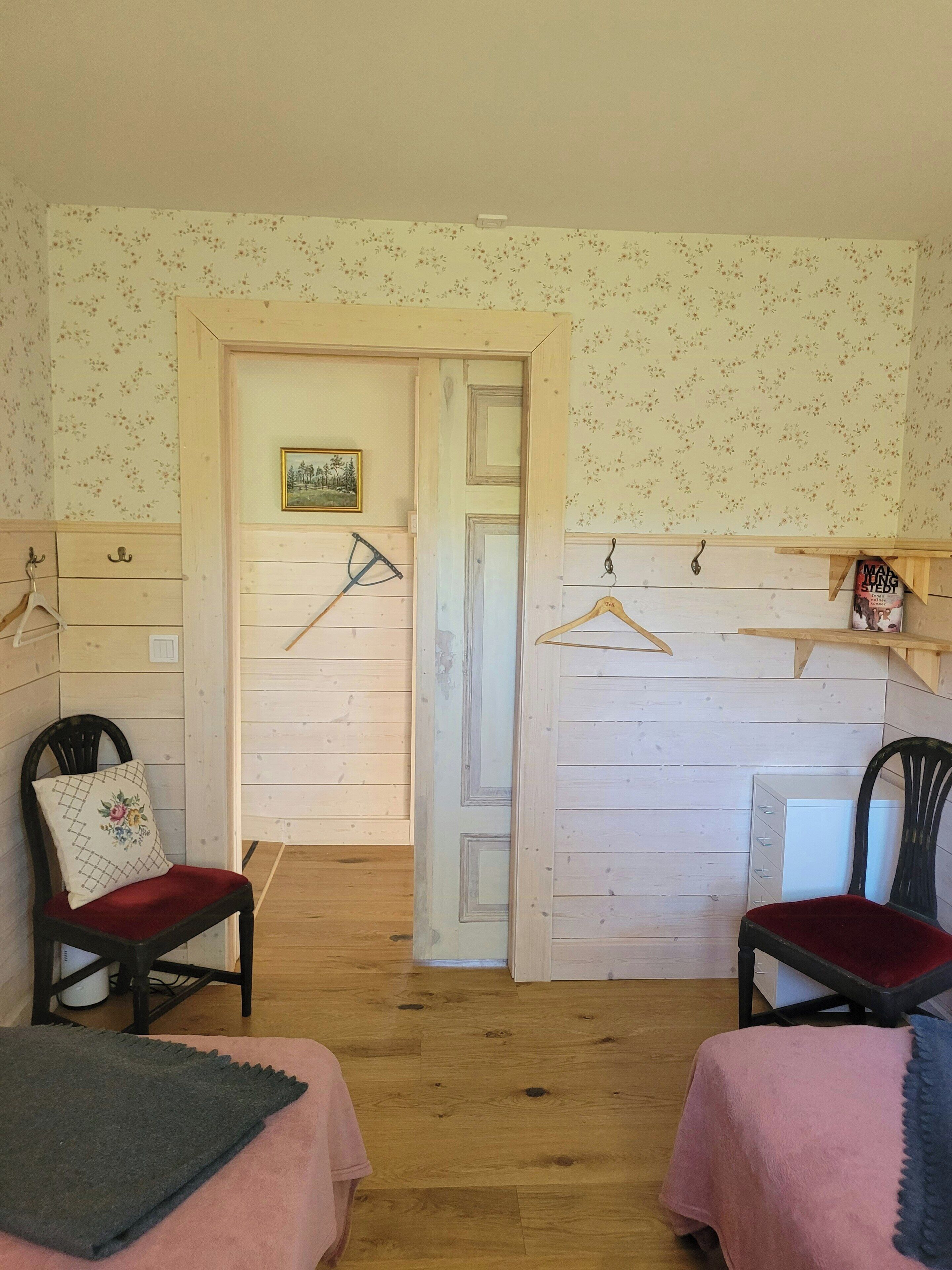 A room with two beds, wooden floors, a door with a picture, and a small shelf with a book.