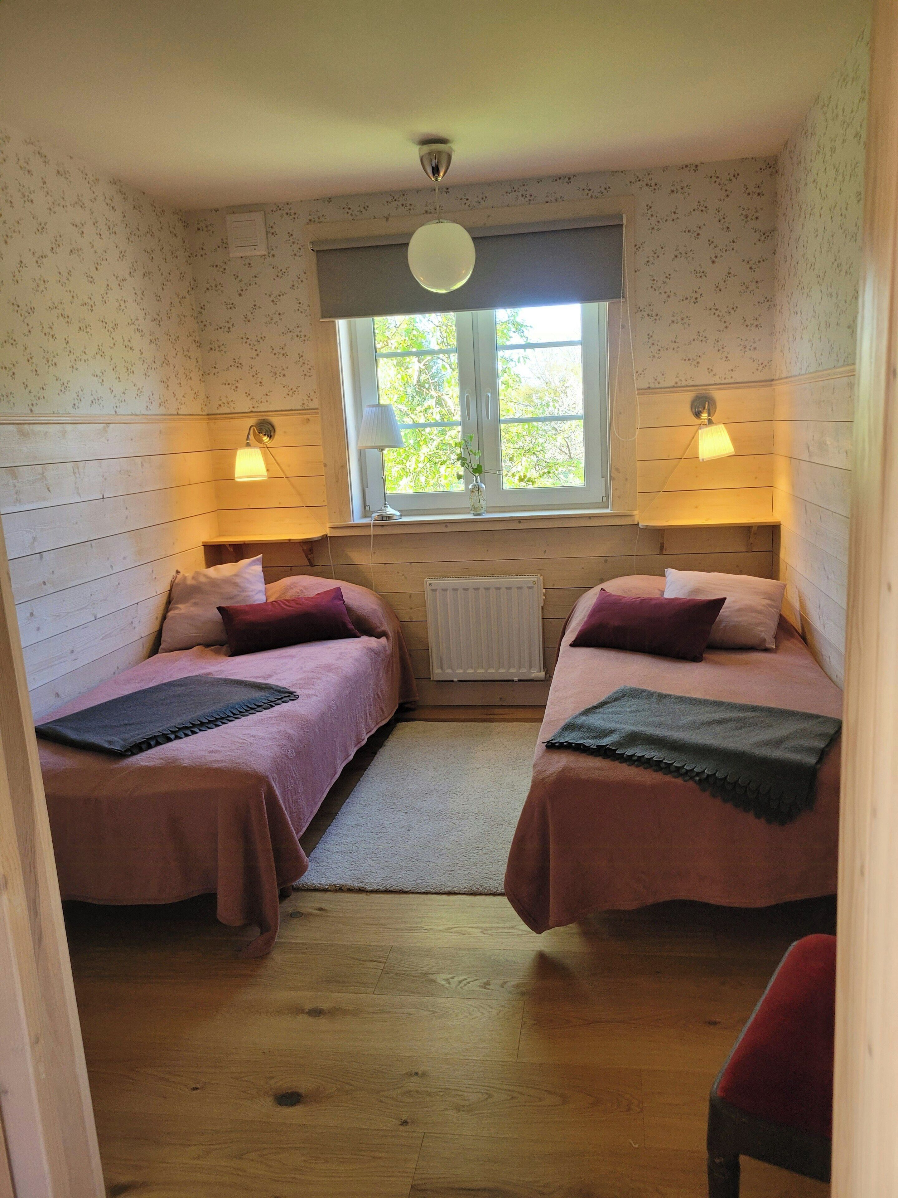 A room with two beds, a window with a view, a radiator, and two wall-mounted lamps.