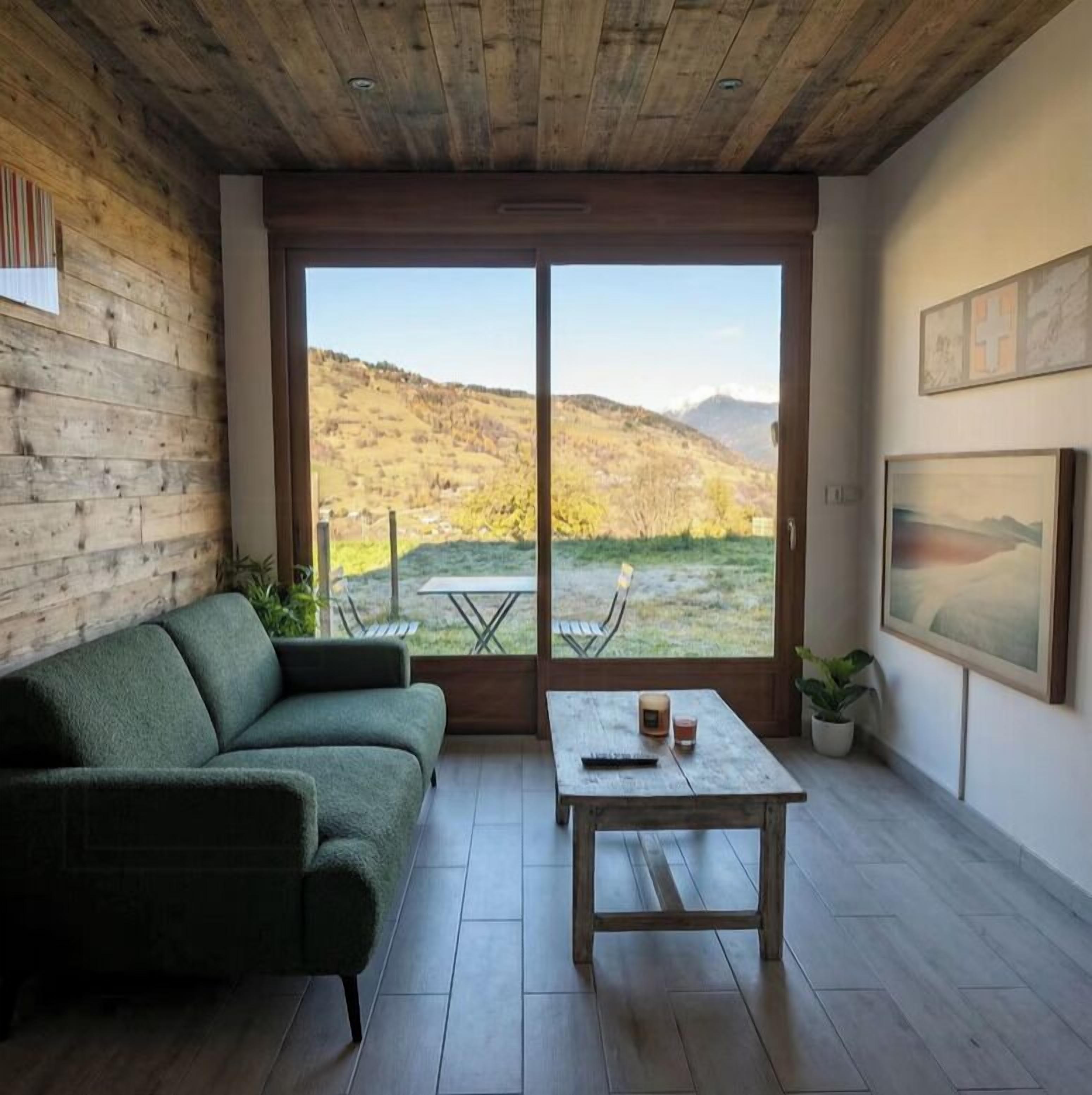 Una sala moderna con un sofá verde, una mesa de centro de madera y un amplio ventanal corredero que da a un área exterior con vistas a las colinas.