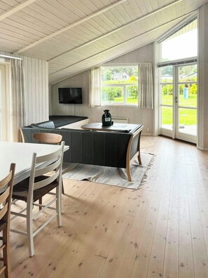 Un salon moderne comprenant un canapé, une table à manger avec des chaises, un téléviseur fixé au mur et de grandes fenêtres offrant une vue sur l’extérieur.