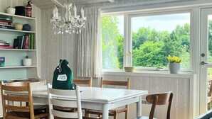 A dining area with a white table and chairs, a chandelier, a bookshelf with books and decorative items, and a window with a view of greenery. - Family retreat by Roskilde Fjord (Ølsted)