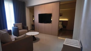 A modern living room with a sofa, a coffee table, a flat-screen TV mounted on a wooden panel, and a small table with a lamp. - Starline Hotel (Dodoma)