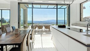 A modern kitchen with a large island, a dining area with chairs, and a view through large windows. - Villa with panoramic views and pool in Gordes (Gordes)