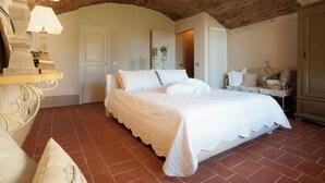 Une chambre avec un plafond en briques, un lit recouvert de draps blancs, une table de chevet avec une lampe et une chaise avec des coussins.