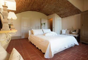 Ein Schlafzimmer mit Ziegeldecke, einem Bett mit weißen Leinen, einem Nachttisch mit Lampe und einem Sessel mit Kissen.